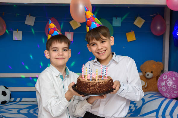 Two boys wearing party hats holding a birthday cake with candles in a decorated room.