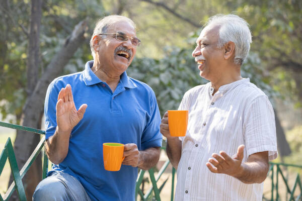 Two older men laughing and talking while holding orange mugs outdoors.
